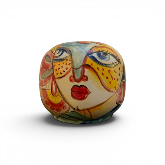 A round ceramic handmade vase like a stylized face, decorated with vibrant hand-painted patterns in yellow, orange, red, blue, and green on a white background.