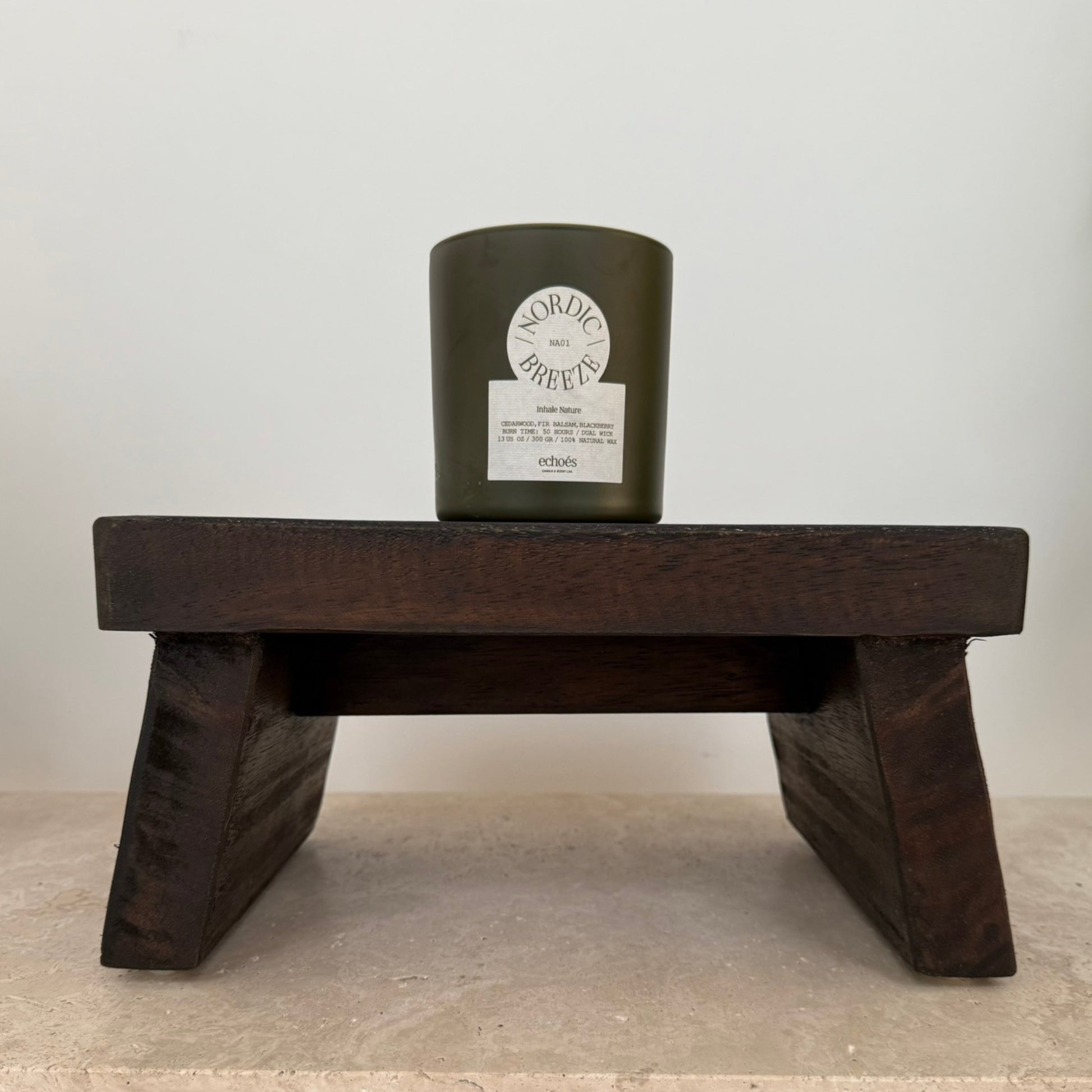 Low aged teak wood styling pedestal with angled slab legs, featuring a textured Echoes Natural candle displayed on top